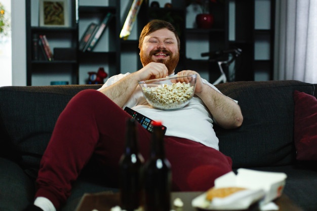 Laughing fat man sits on the sofa, eats pop-corn and watches tv
