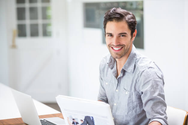 Smiling young man holding documents while sitting at desk with laptop 