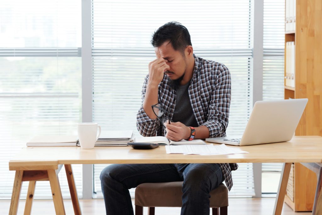 Stressed asian man sitting at table with laptop and documents and rubbing forehead