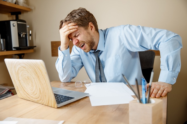 Feeling sick and tired. frustrated sad unhappy sick young man massaging his head while sitting at his working place in office