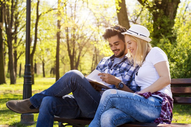 Happy caucasian couple sitting on a park bench, reading a book together