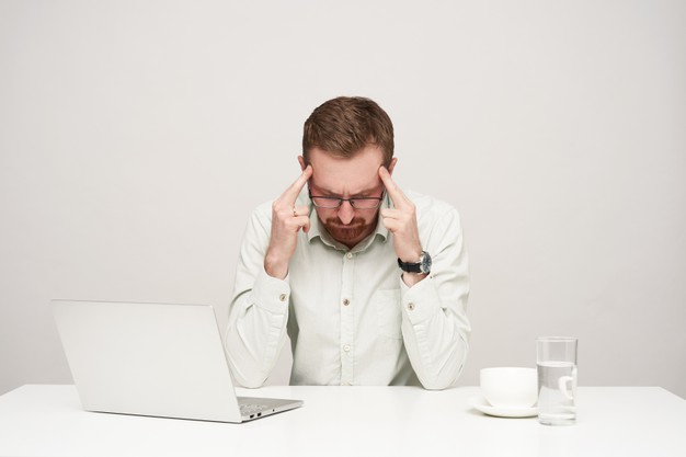 Tired young unshaved fair-haired businessman keeping fingers on temples while having headache after hard working day