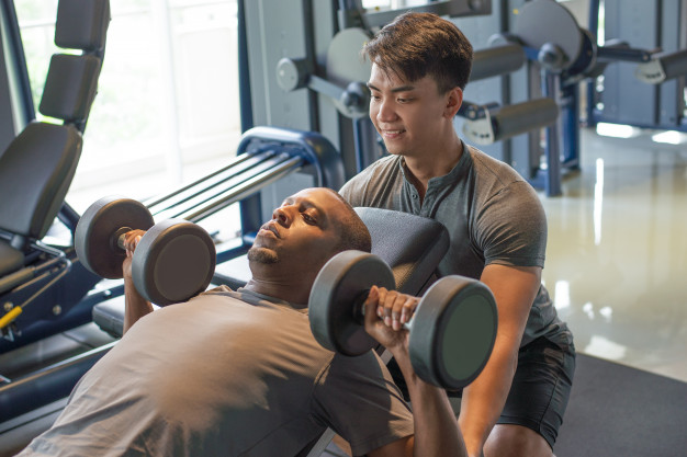 Trainer and man lying and lifting dumbbells in gym 
