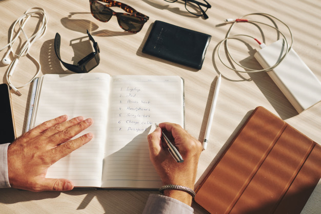 unrecognizable-man-writing-plan-journal-gadgets-lying-around-desk