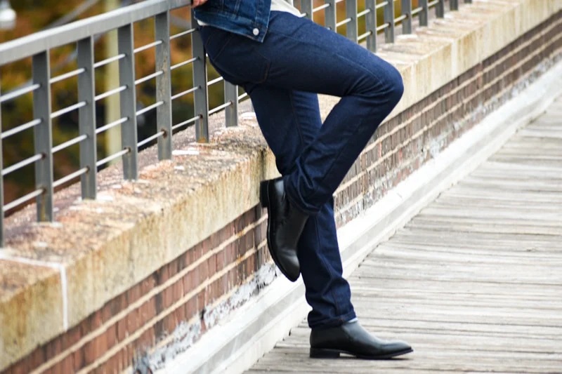 Best-Jeans-for-Men-Model-Outside-Leaning-Against-Guard-Rail-Wearing-Slim-Fit-Dark-Wash-Jeans-and-Black-Boots