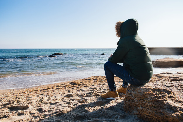 man sitting on the stone, looking at the sea. dressed in warm jacket with hood