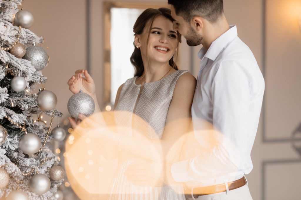 Fancy dressed man and woman in silver gown hug each other tender standing before a christmas tree