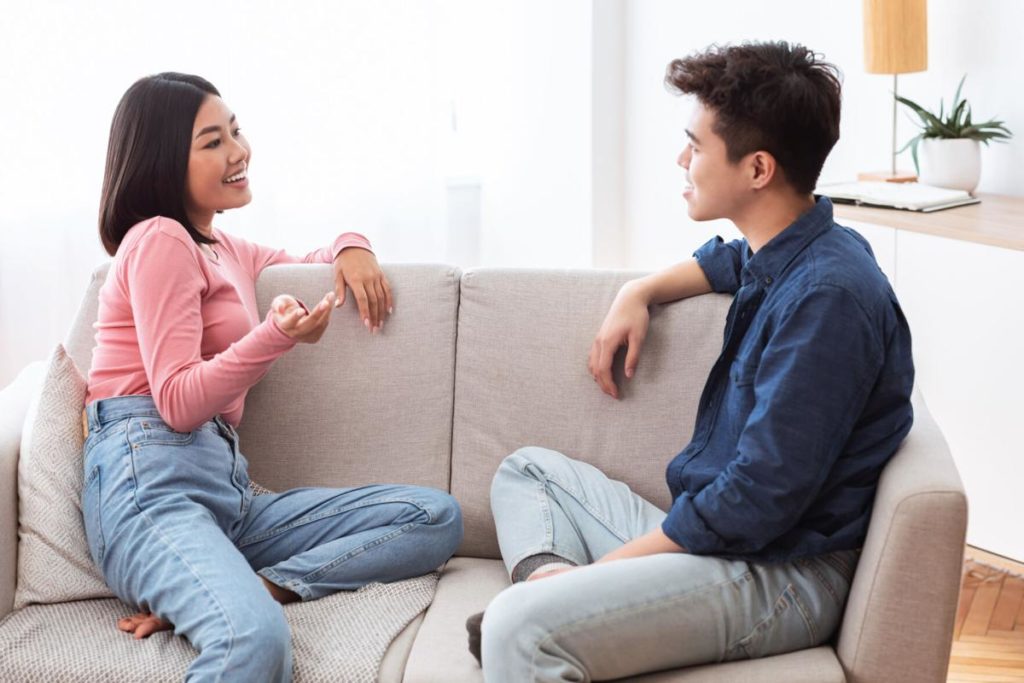 Asian Couple Talking Enjoying Conversation Sitting On Couch At Home