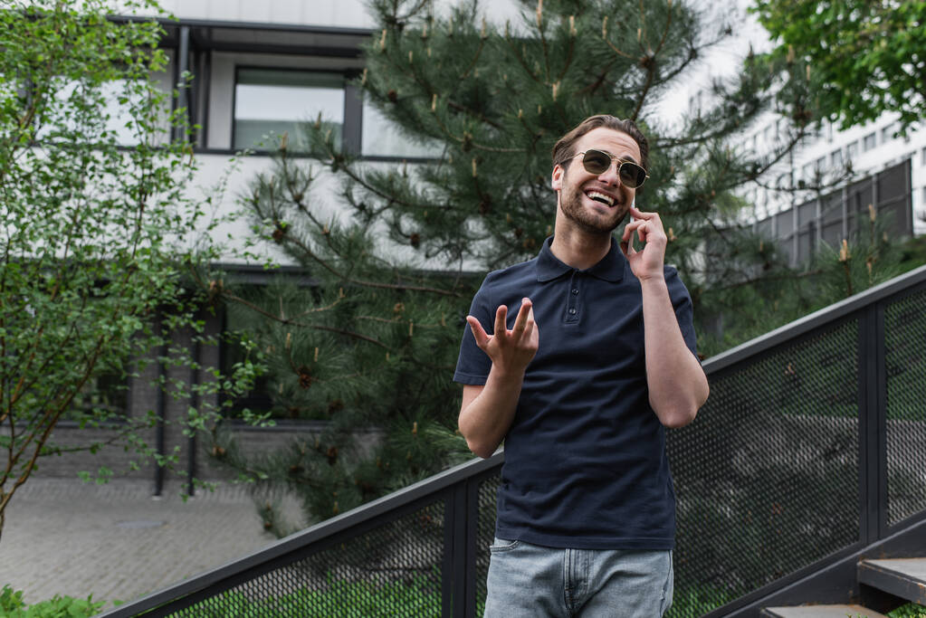 Cheerful man in sunglasses and polo shirt talking on cellphone outside