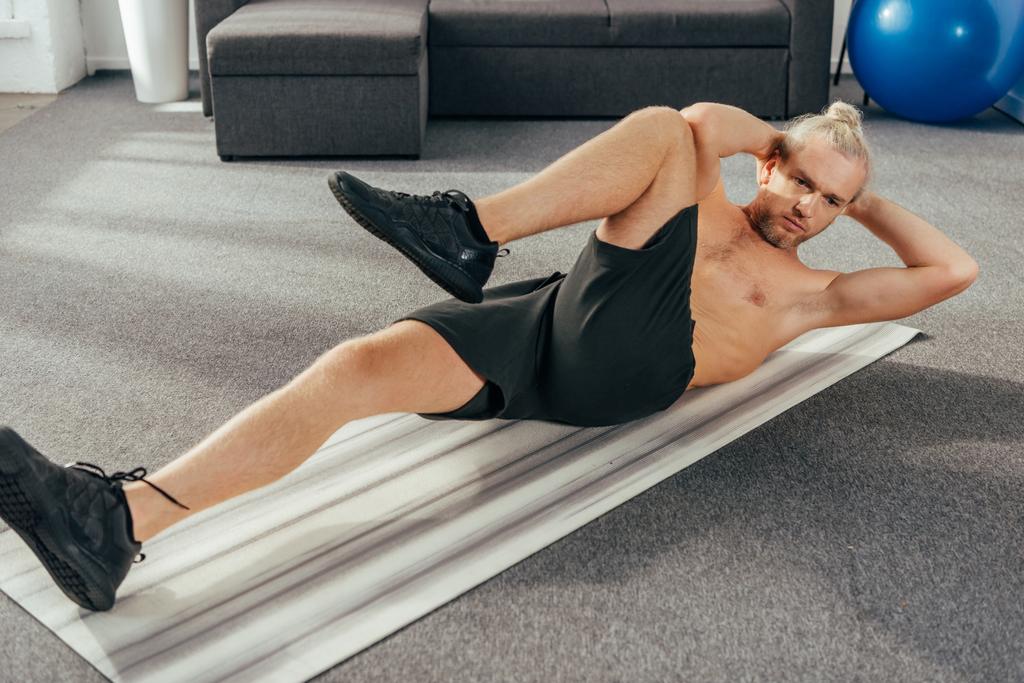 Muscular shirtless man doing abs exercise on yoga mat at home