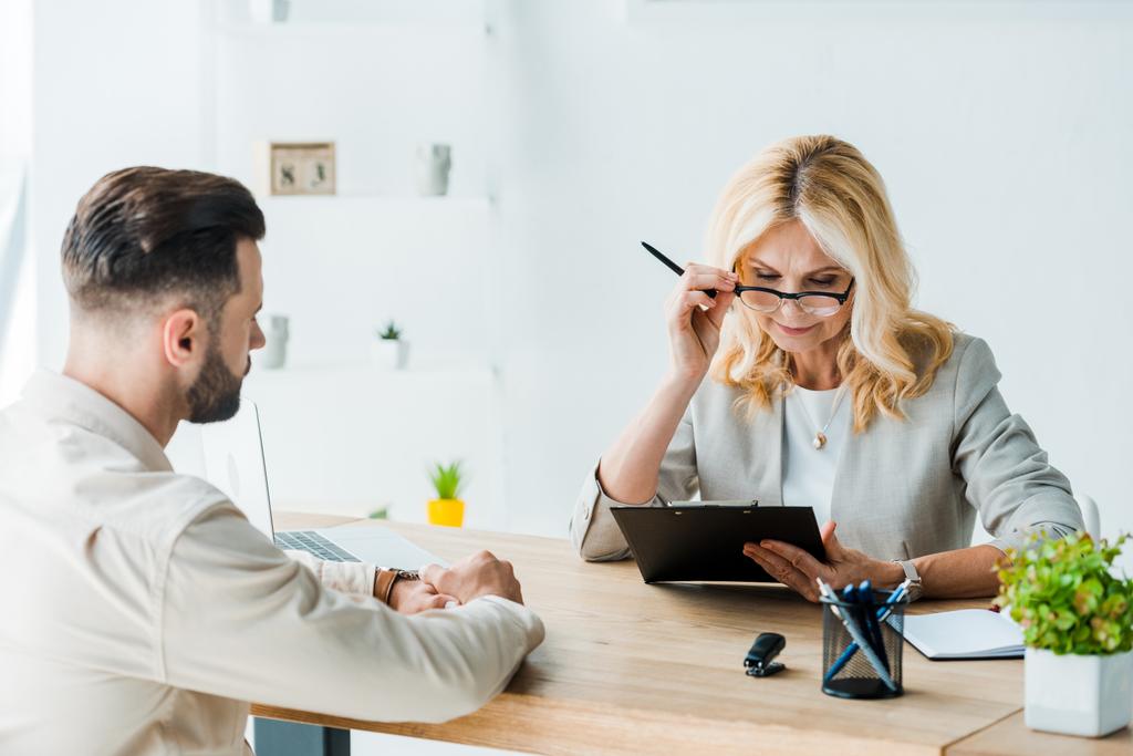 blonde recruiter in glasses looking at clipboard near bearded man 