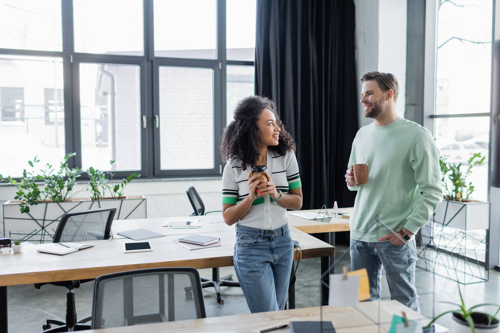 Smiling interracial business colleagues holding coffee while talking in office