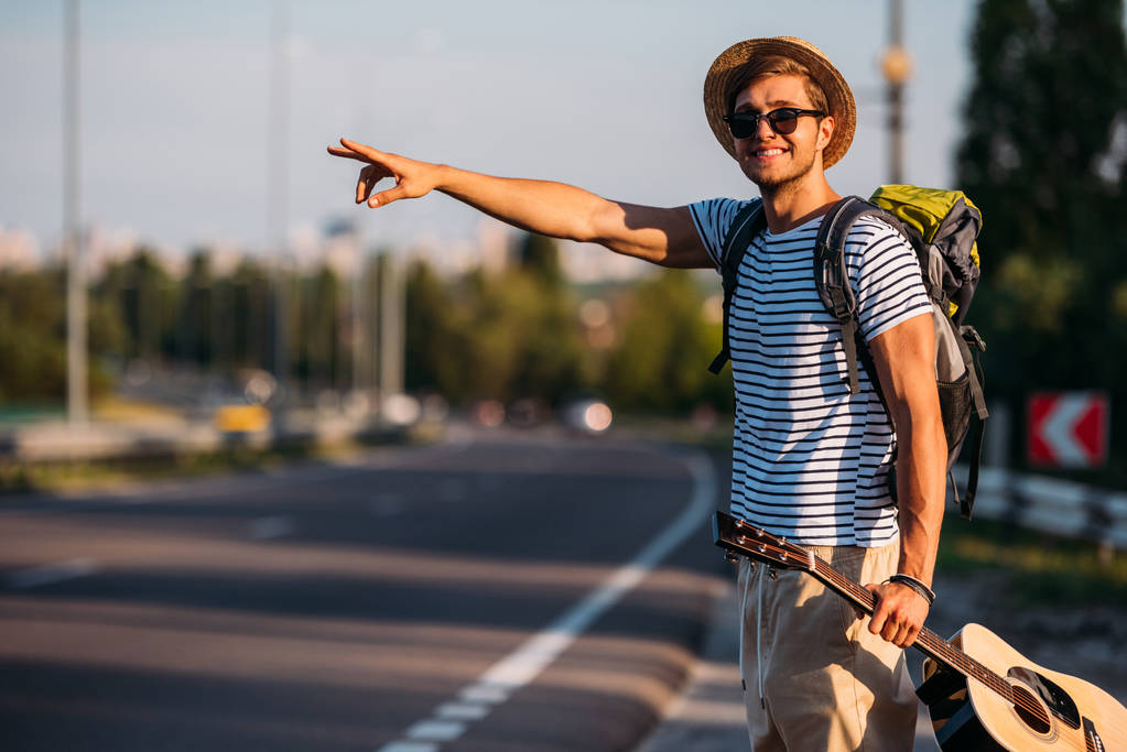 Smiling man with backpack and guitar in hand gesturing to stop car while hitchhiking