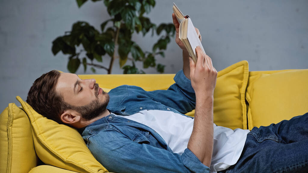 man lying on sofa and reading book in living room
