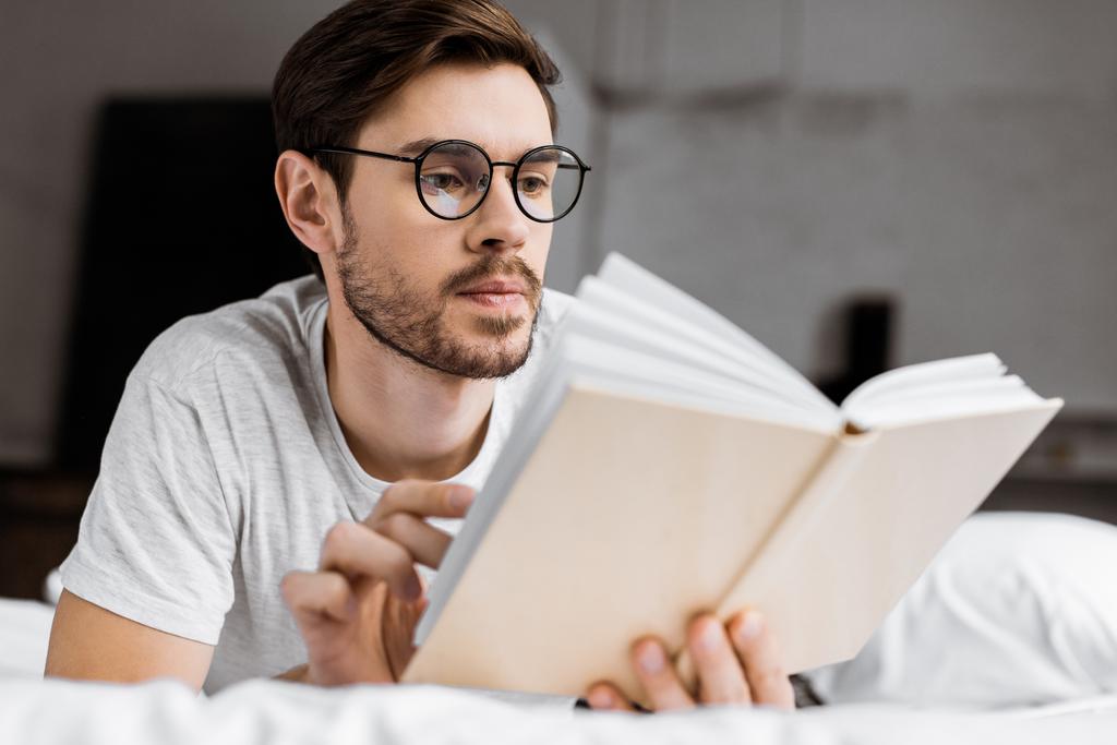 young man in eyeglasses lying in bed and reading book