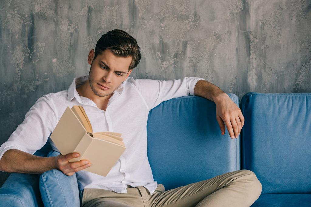 man reading book while sitting on sofa
