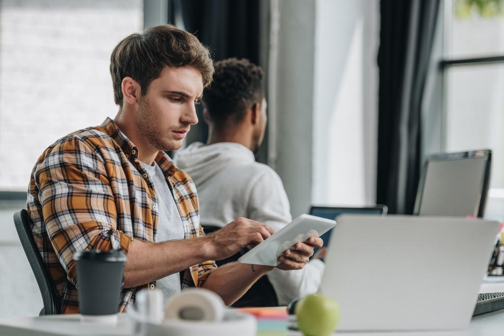 programmer using digital tablet while sitting at workplace near african american colleague