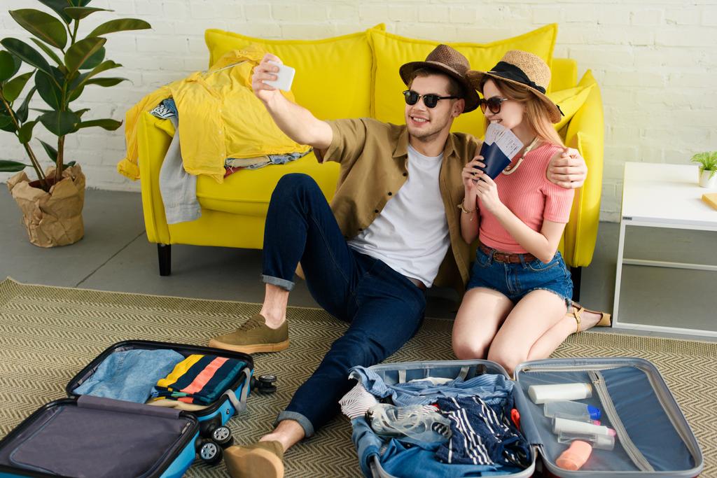 couple taking selfie on smartphone while packing suitcases for vacation 