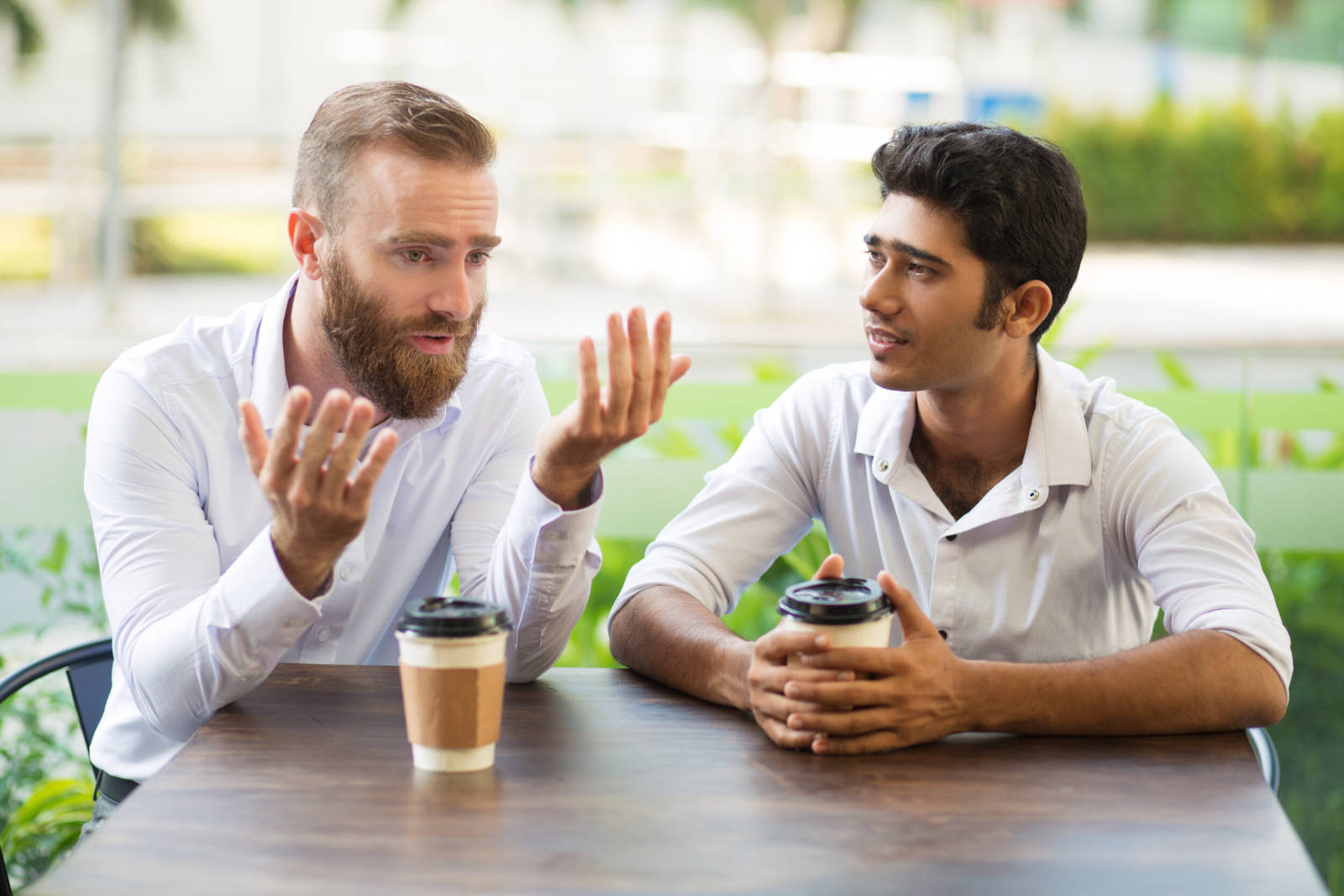 Two male friends drinking coffee and talking in outdoor cafe. People sitting at table with blurred view in background. Coffee break