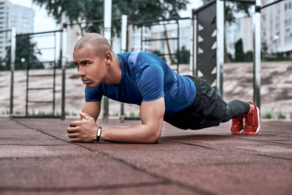 Handsome african man is doing stretching exercises at open air gym near the park. He uses exercises for his abdomen adn doing plank