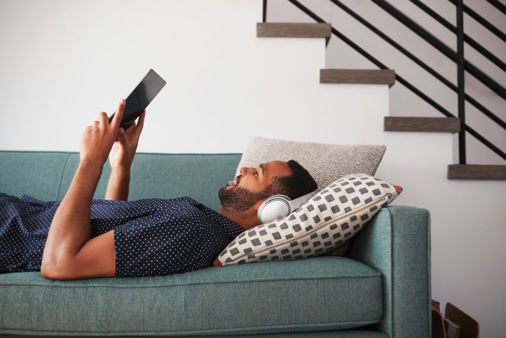 Man Lying On Sofa At Home Wearing Headphones And Watching Movie On Digital Tablet