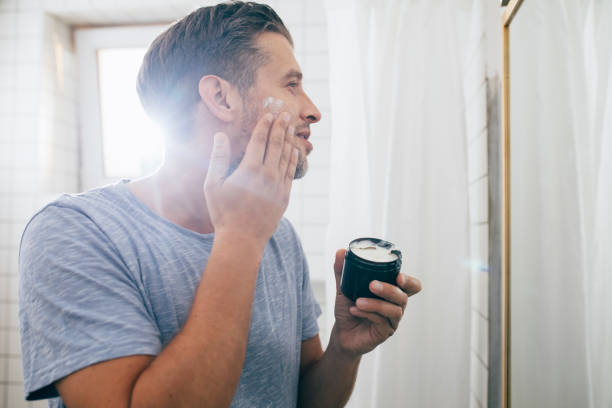 Handsome happy young Caucasian man applying face cream after a morning shave