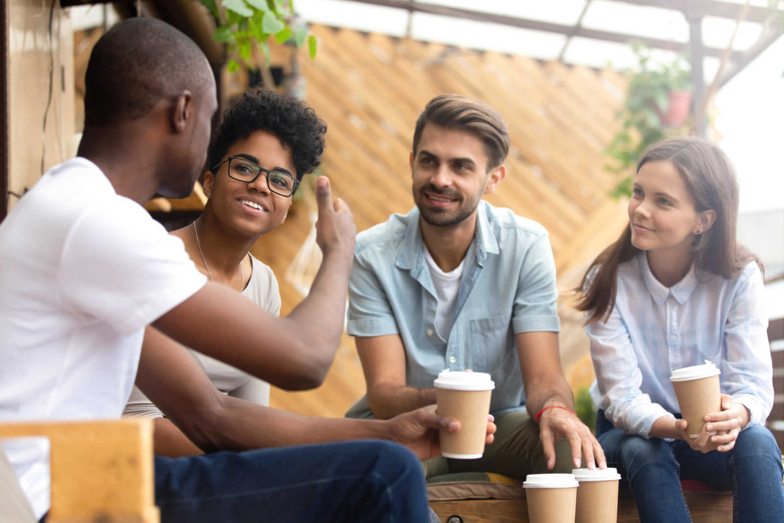 African American man showing thumb up, telling story to interested attentive friends, sitting, drinking coffee in cafe together, smiling multiethnic friends discussing, talking, chatting