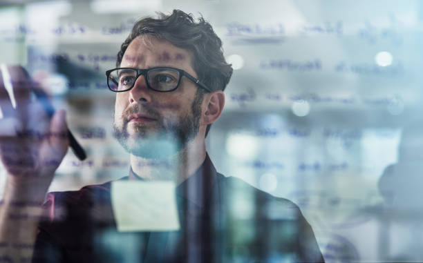 Shot of a young businessman brainstorming with sticky notes on a glass wall in a modern office