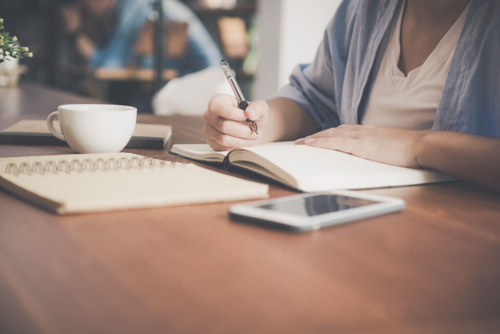 Young business woman in white dress sitting at table in cafe and writing in notebook. Asian woman talking smartphone and cup of coffee. Freelancer working in coffee shop. Vintage effect style pictures
