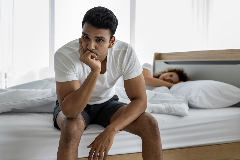Upset depressed young man sitting on the edge of bed against his wife lying on the bed.